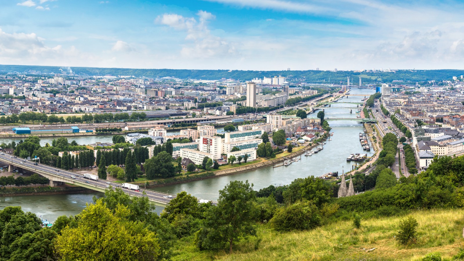 Vue panoramique de Rouen sur la Seine, ses ponts et le paysage urbain verdoyant sous un ciel bleu.